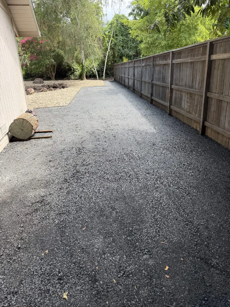 Wide crushed black rock side yard driveway alongside a wood fence